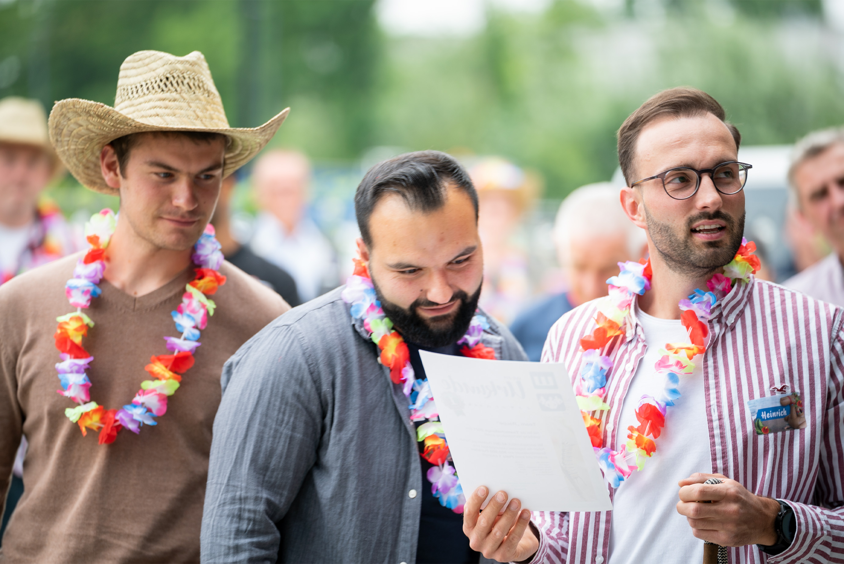 Zwei Männer mit Hawaiikette schauen auf ein Blatt Papier, ein anderer hält es in der Hand und schaut woanders hin