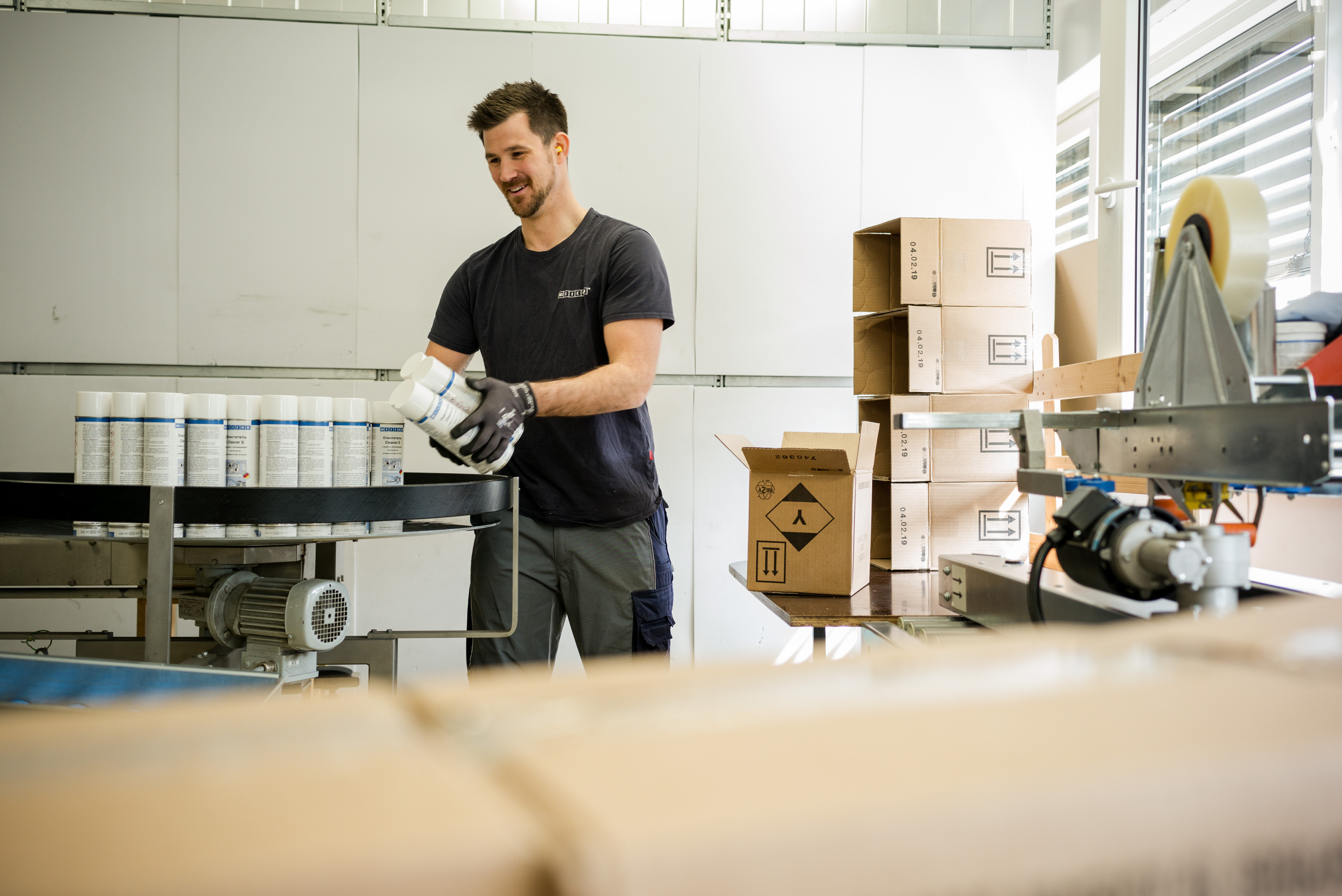 Ein Mann arbeitet iim Lager und hält mehrere Spraydosen in der Hand, weitere stehen auf einem Fließband