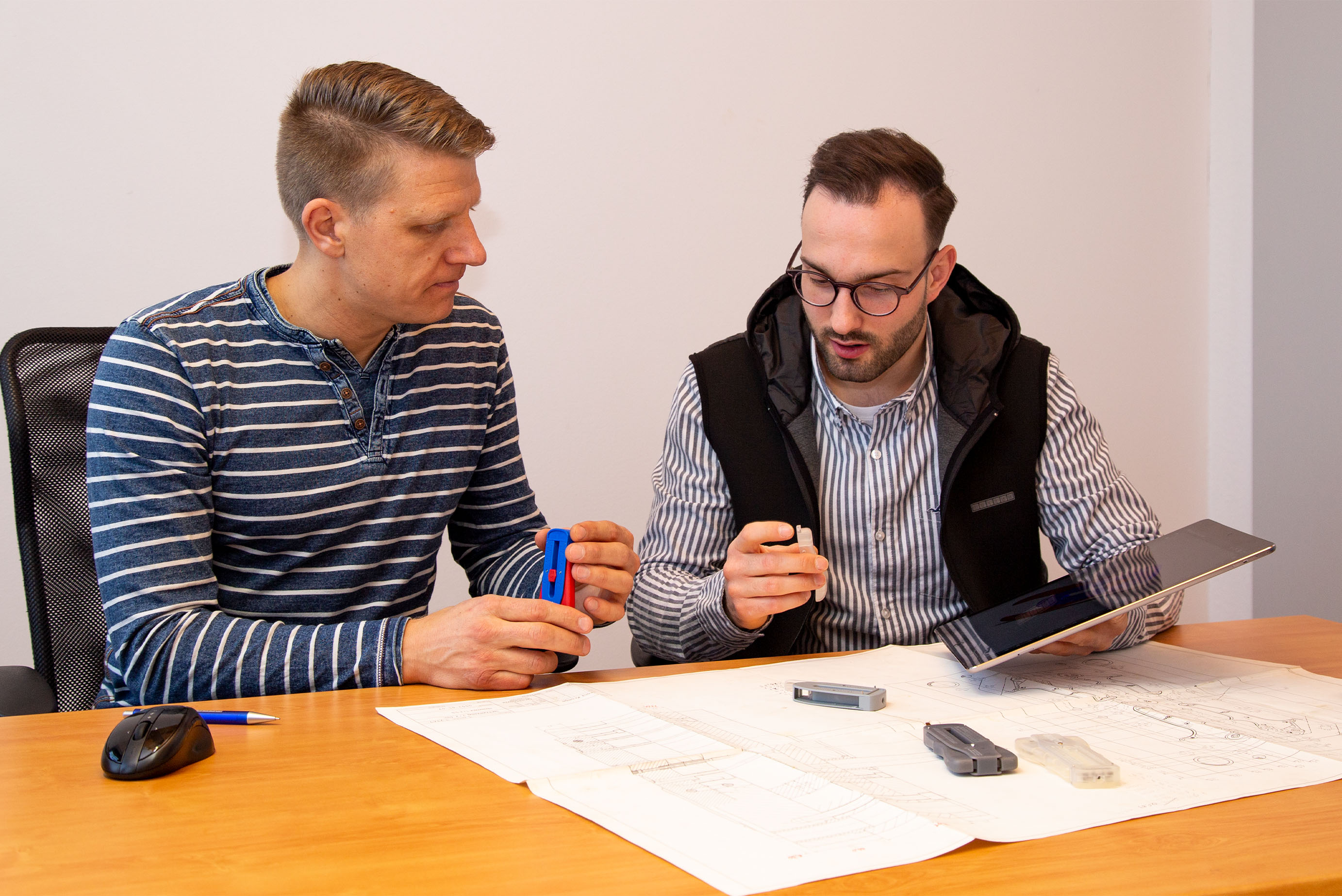 Zwei Männer sitzen an einem Schreibtisch und schauen sich einen Plan an, einer hat ein Werkzeug in der Hand, der andere ein Tablet