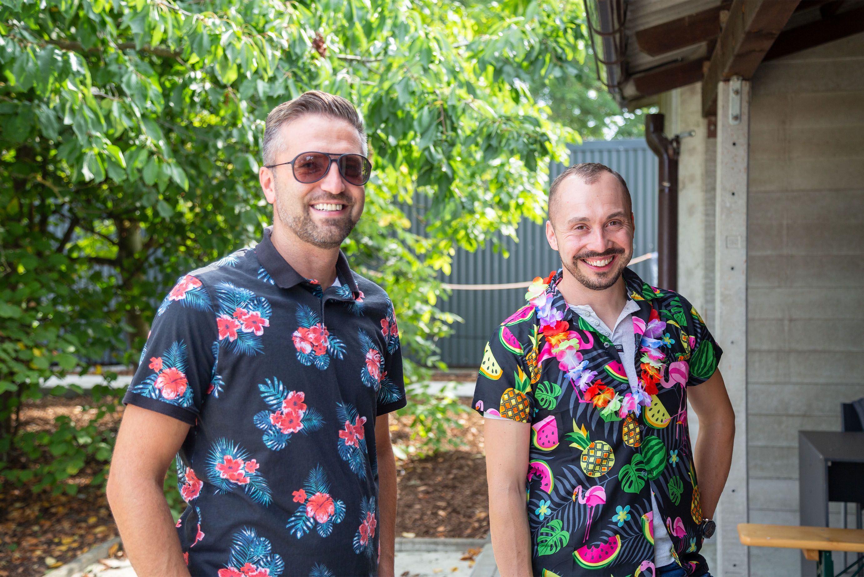 Zwei Männer mit Hawaiihemden stehen an einem Schuppen und lachen in die Kamera