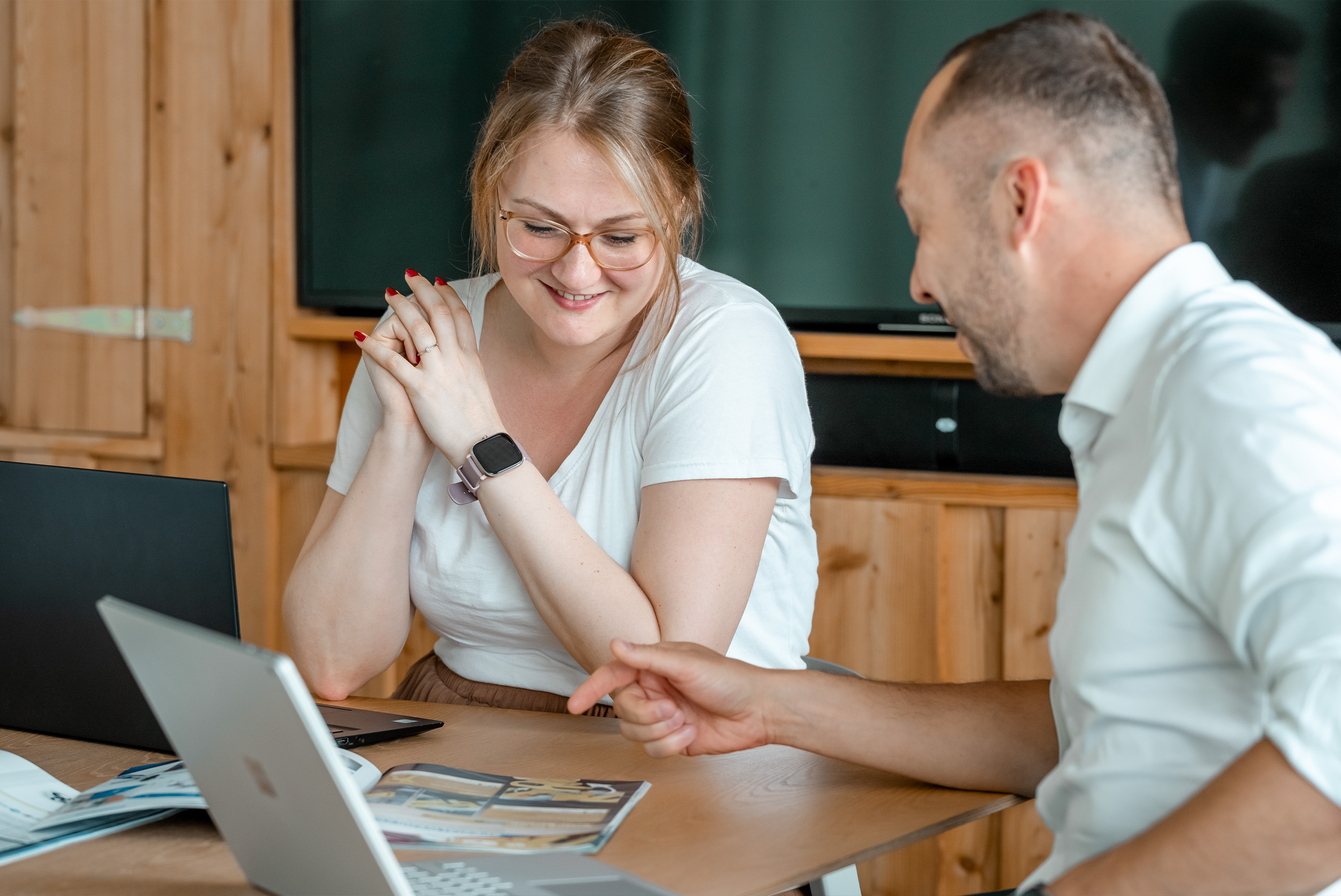 Eine Frau und ein Mann sitzen in einem Besprechungsraum am Schreibtisch und schauen in einen Flyer, vor ihnen steht ein Laptop