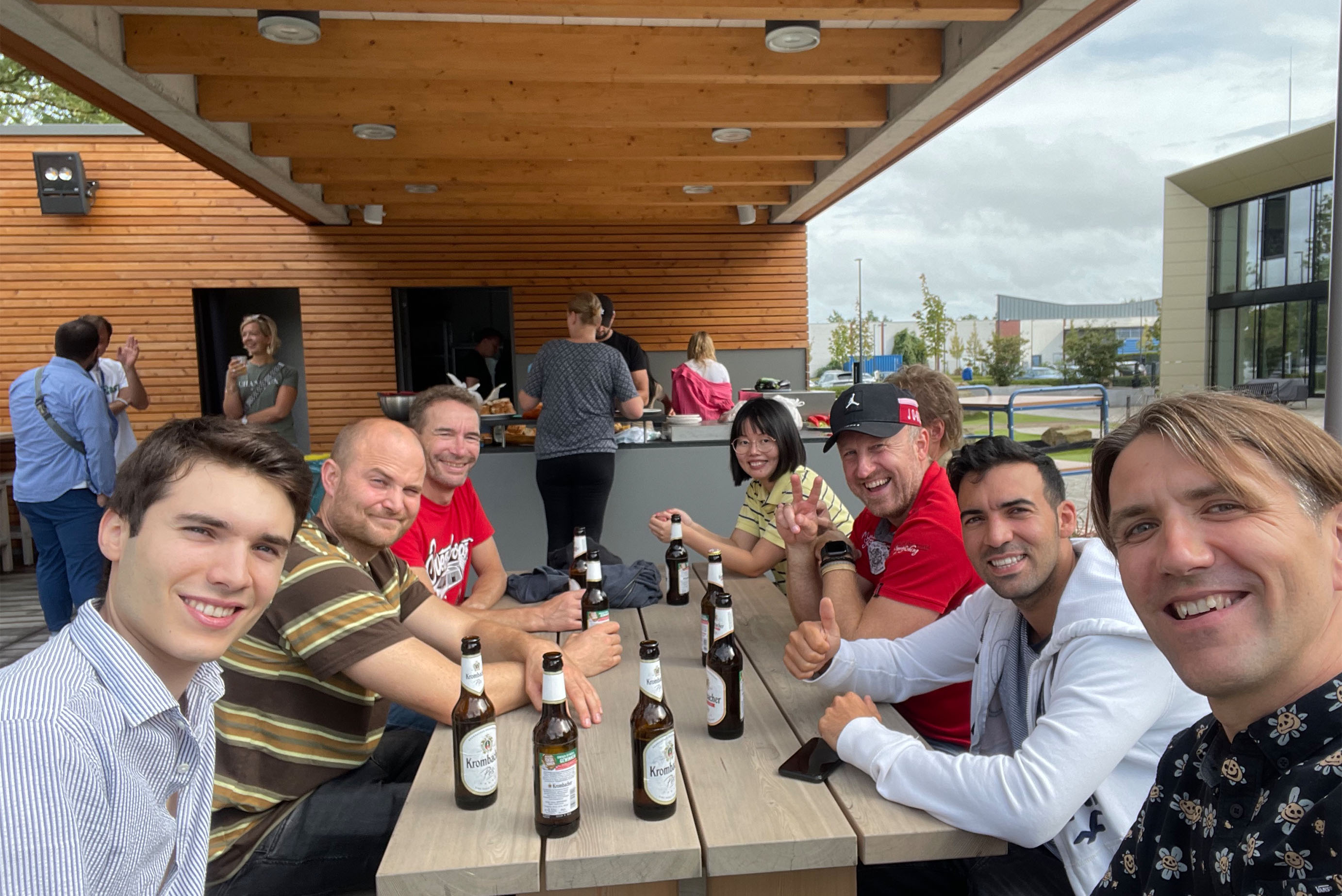 mehrere Personen sitzen auf einer Terrasse an einem Tisch und lachen in die Kamera, einige andere stehen im Hintergrund und unterhalten sich