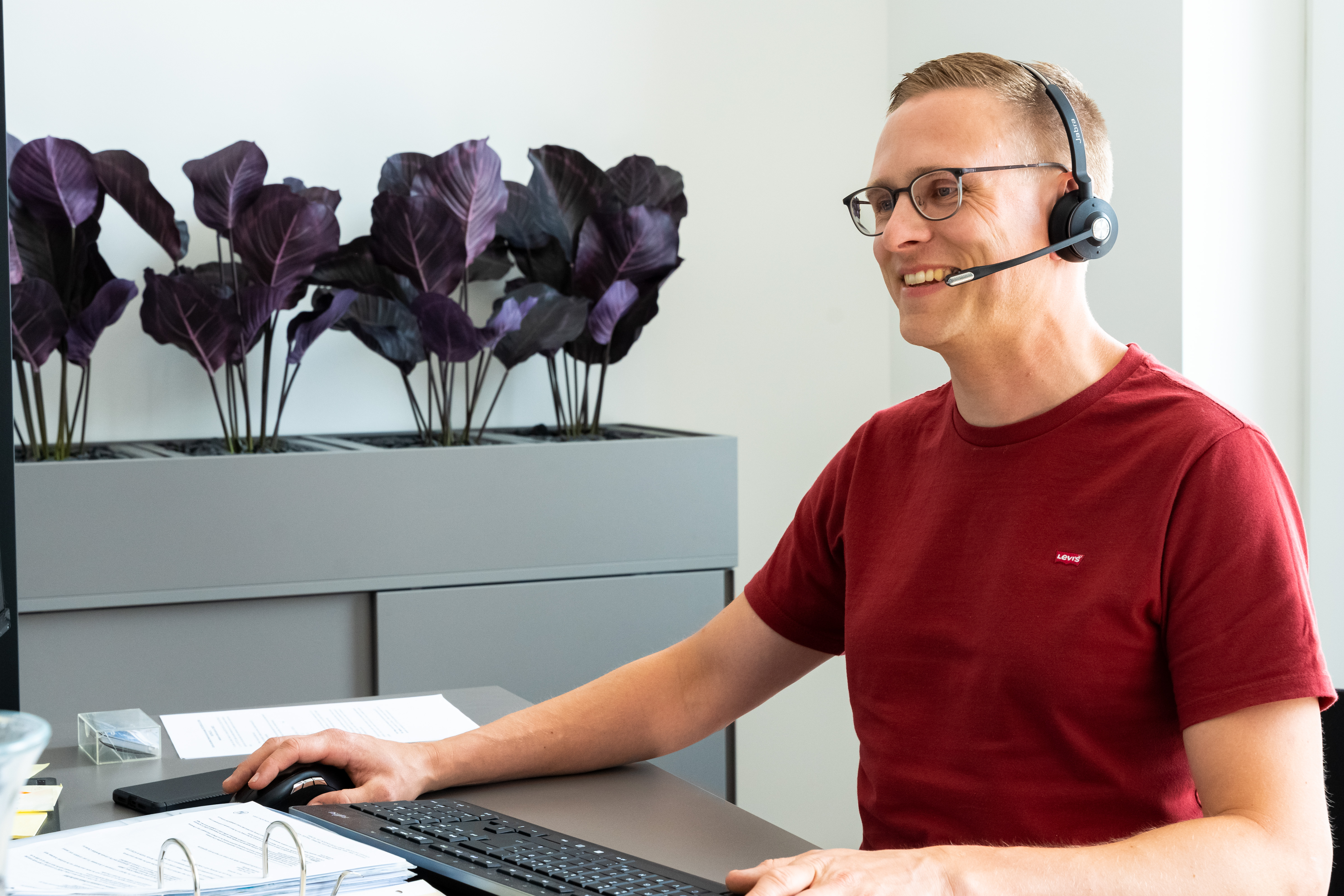 Ein Mann sitzt an einem Schreibtisch, hat ein Headset auf und lacht