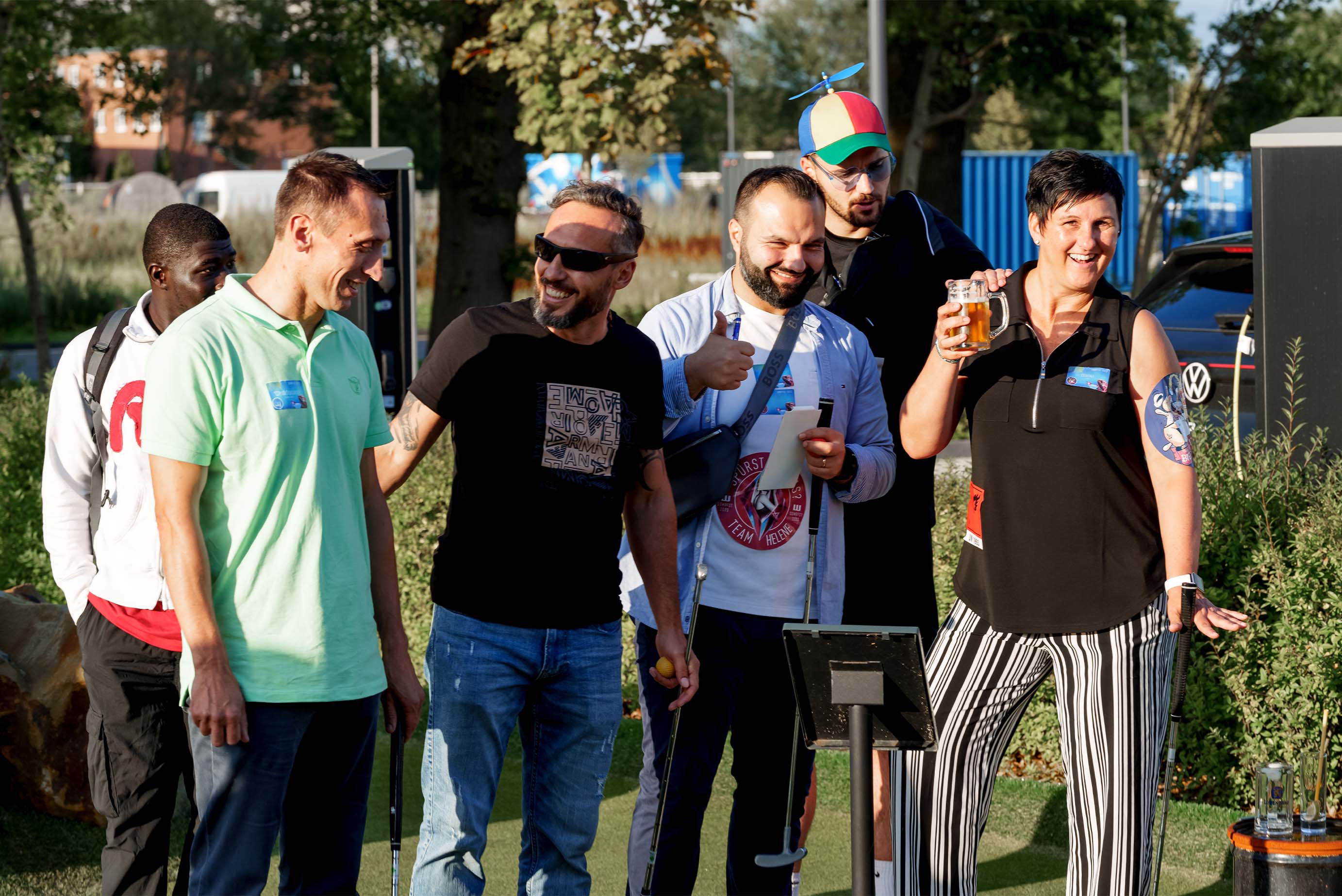 eine kleine Gruppe steht draußen an einem Minigolfplatz und unterhält sich, 2 lachen in die Kamera