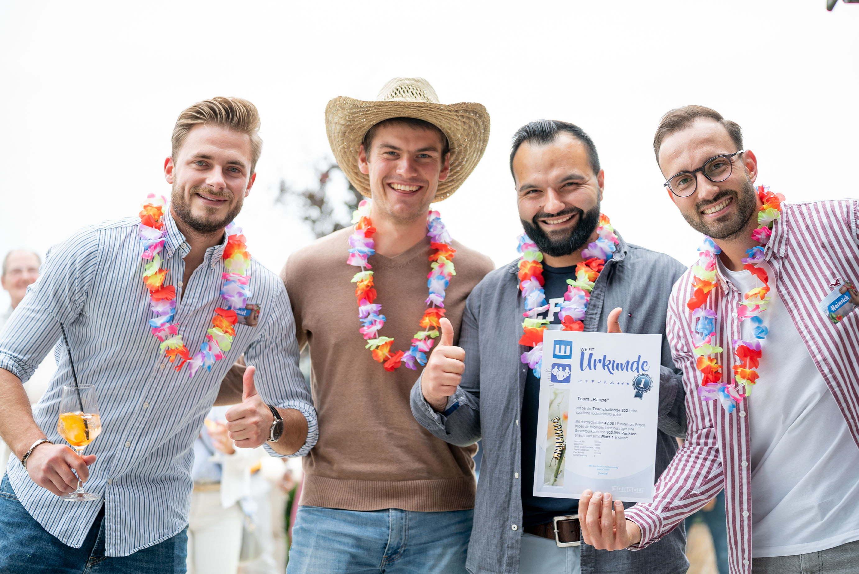 Vier Männer stehe nebeneinander und lachen in die Kamera, sie tragen Hawaiiketten, einer hält ein Getränk in der Hand, ein anderer eine Urkunde