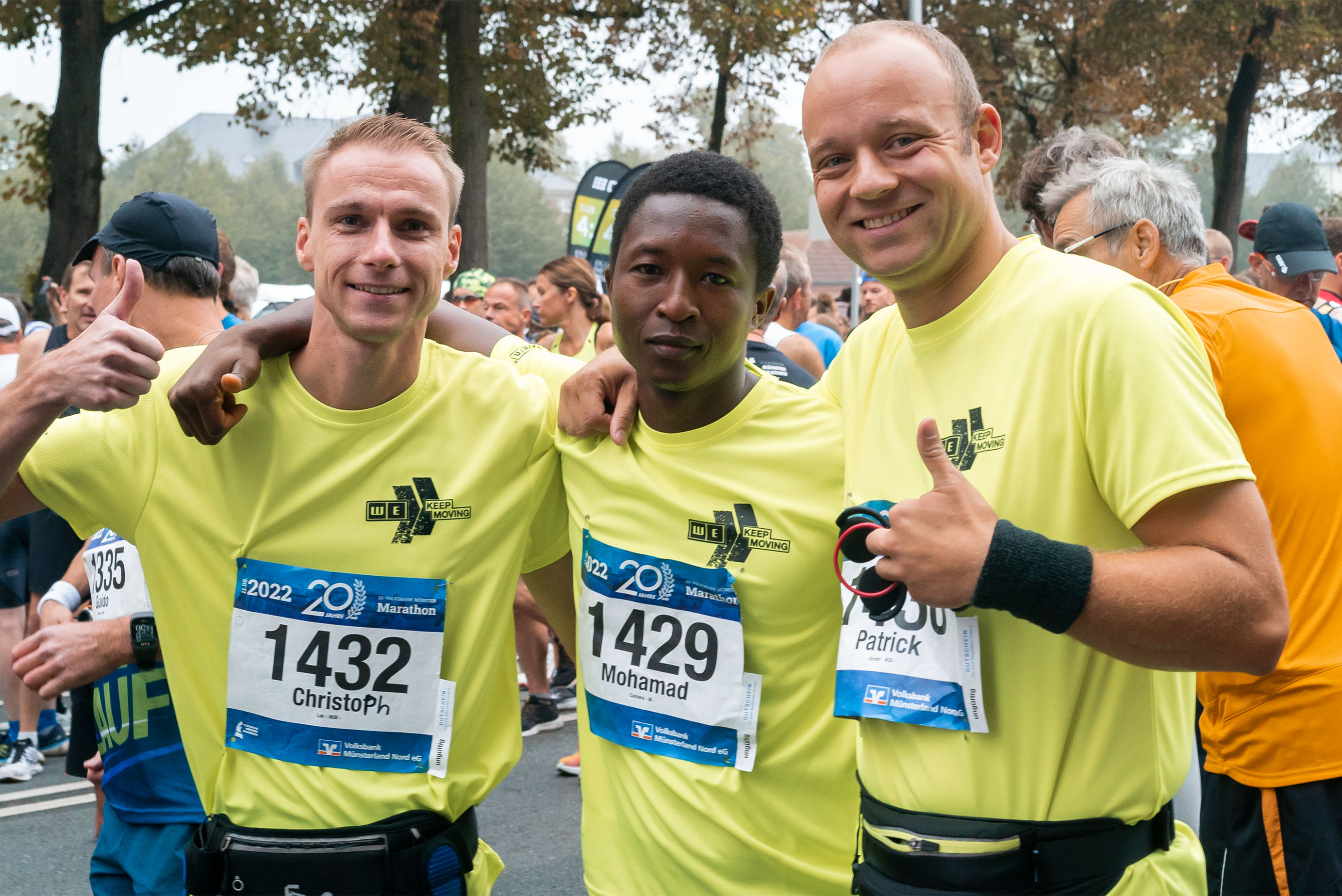 Drei Männer mit gelben Laufshirts und Nummern drauf stehen nebeneinander und lachen in die Kamera