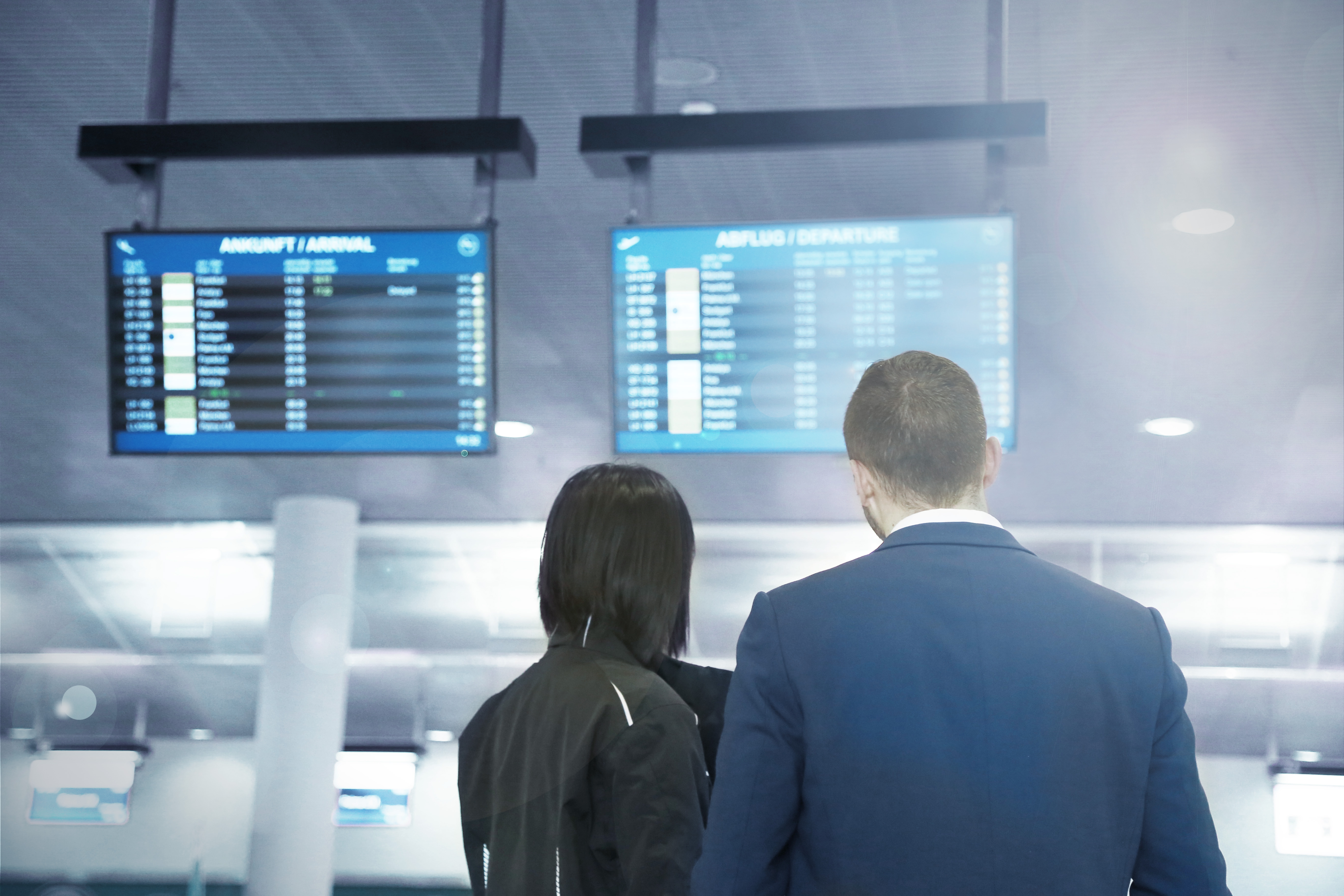 Ein Mann und eine Frau stehen mit dem Rücken zur Kamera an einem Flughafen und schauen auf Bildschirme