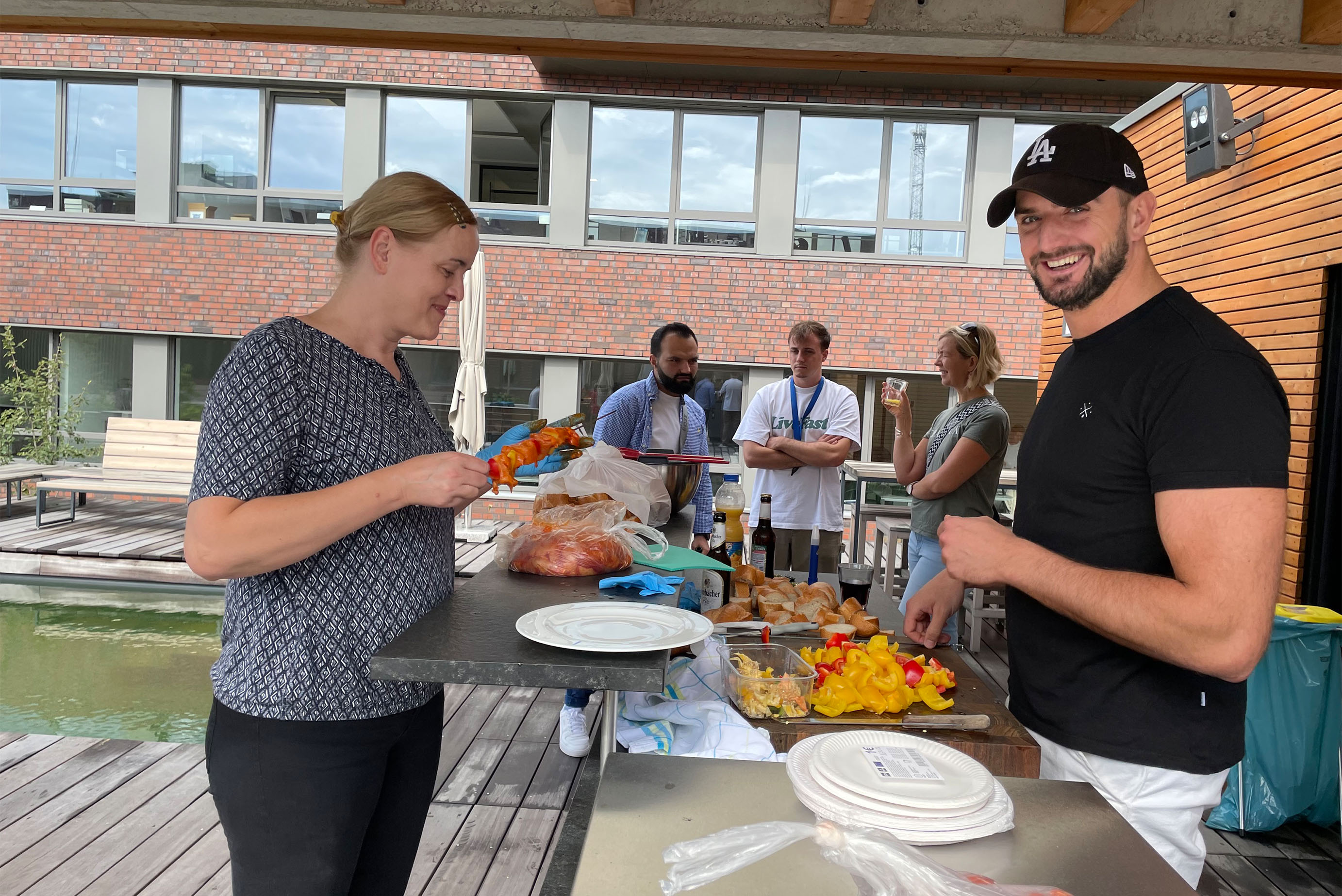 zwei Personen stehen an einer Outdoor-Küche und bereiten Essen vor, im Hintergrund sieht man einen Naturteich und PErsonen die sich unterhalten