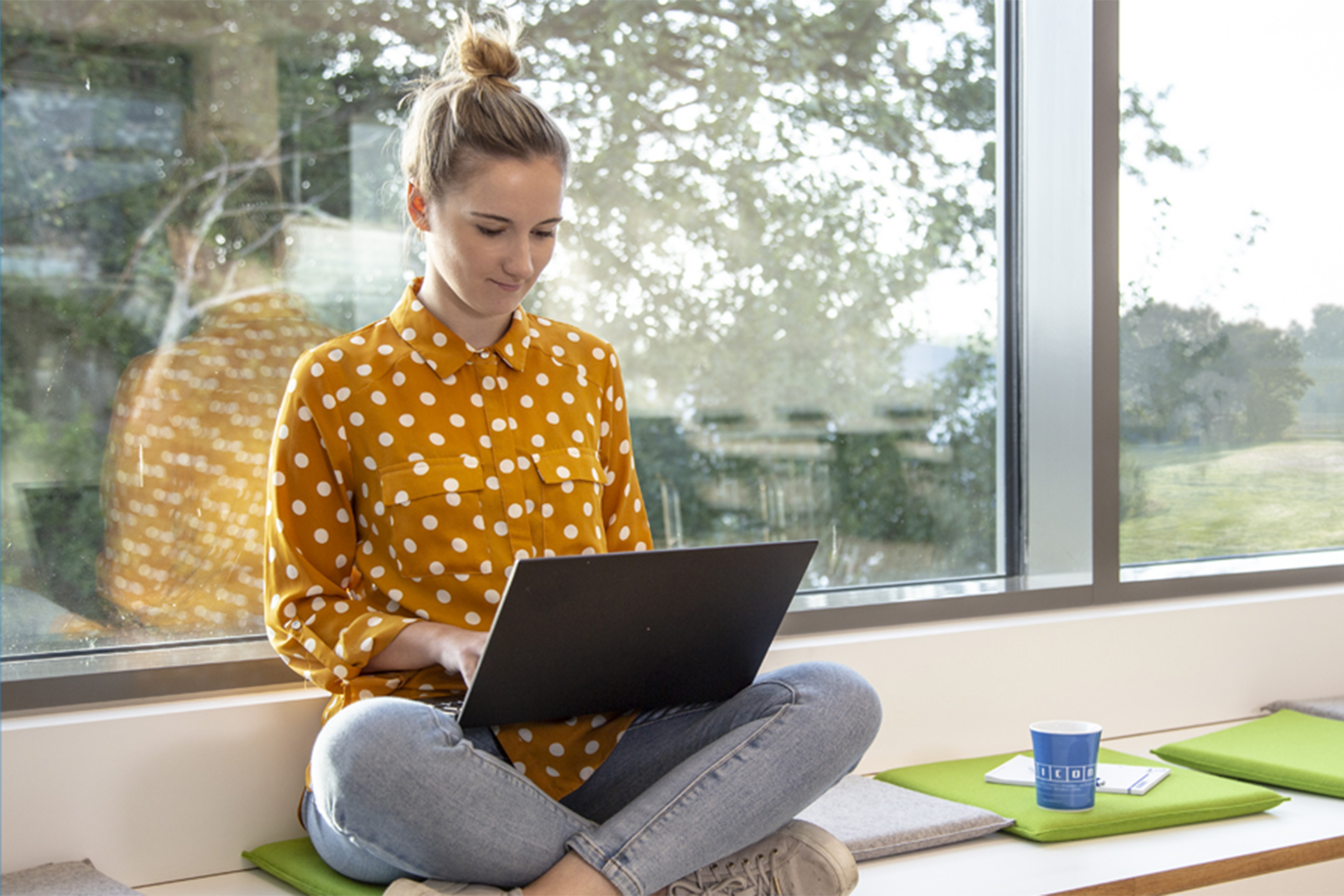 eine Frau sitzt auf einer Fensterbank und hat einen Laptop auf dem Schoß und arbeitet