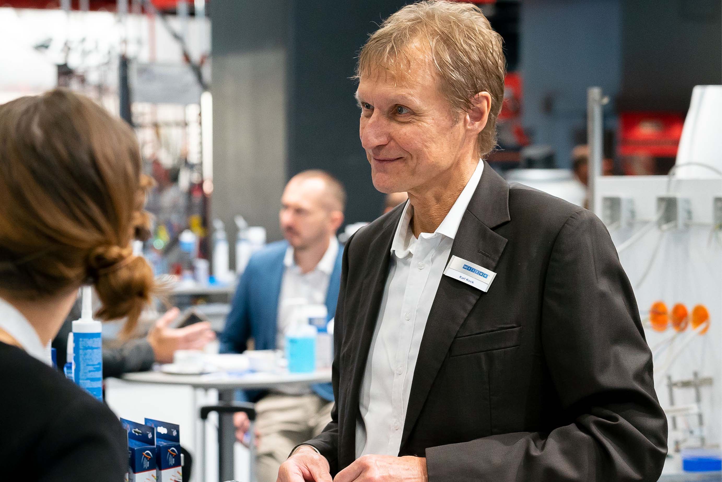 Ein Mann steht mit Anzug an einem Messestand und lächelt jemanden an