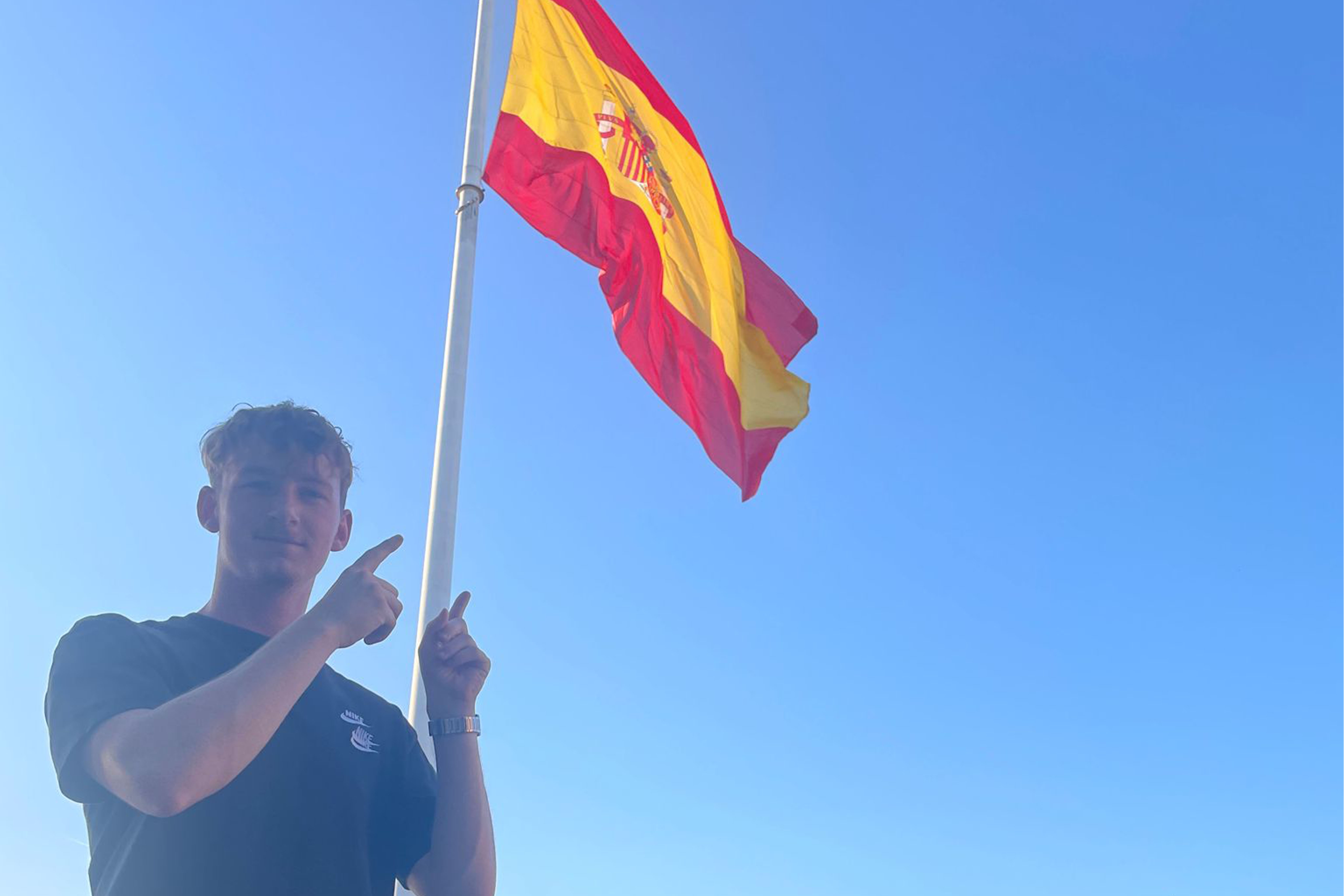 Ein Mann steht neben einer Spanienflagge und zeigt mit beiden Fingern drauf