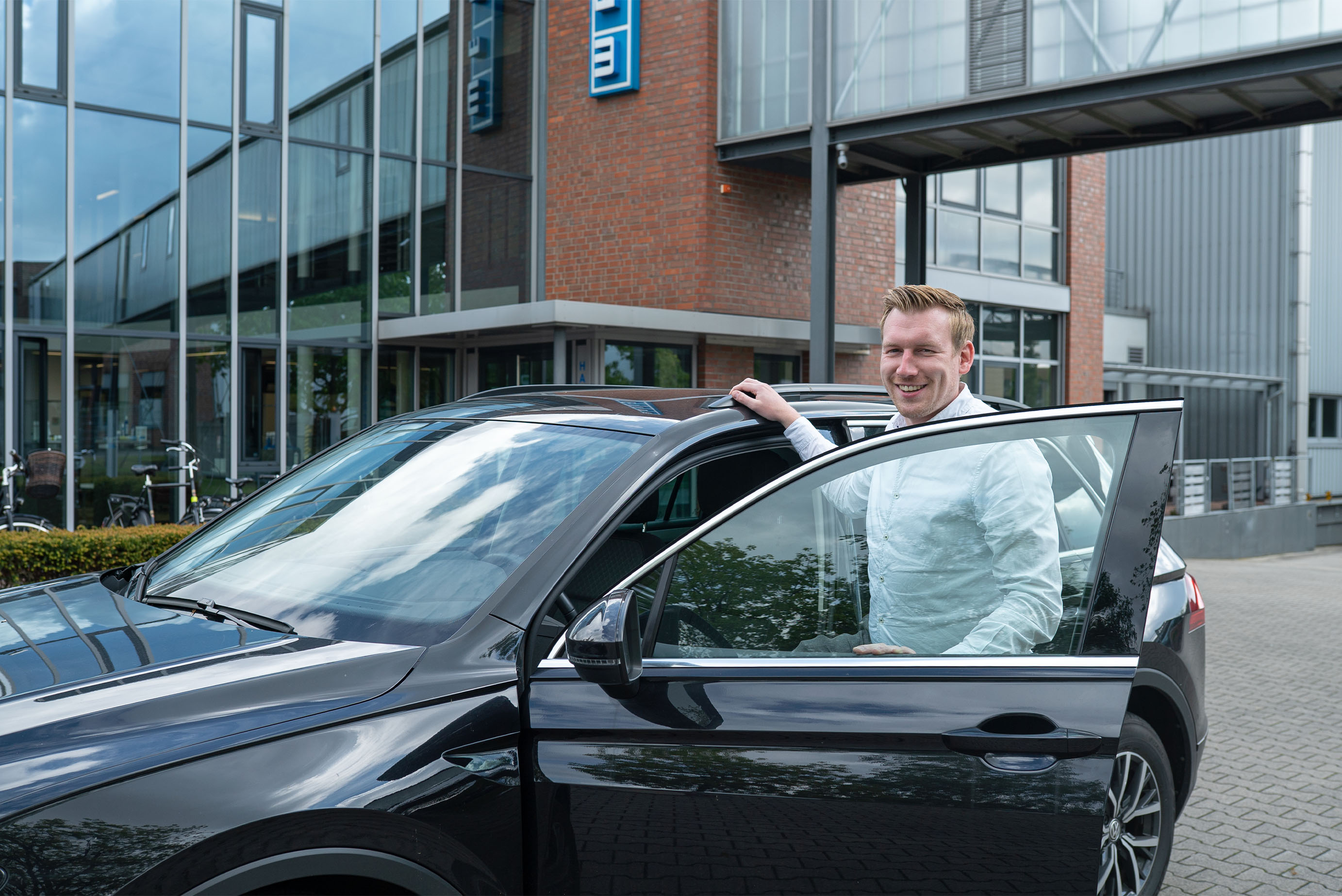 ein Mann mit weißem Hemd steht in einer offenen Autotür eines schwarzen SUVs auf einem Firmengelände