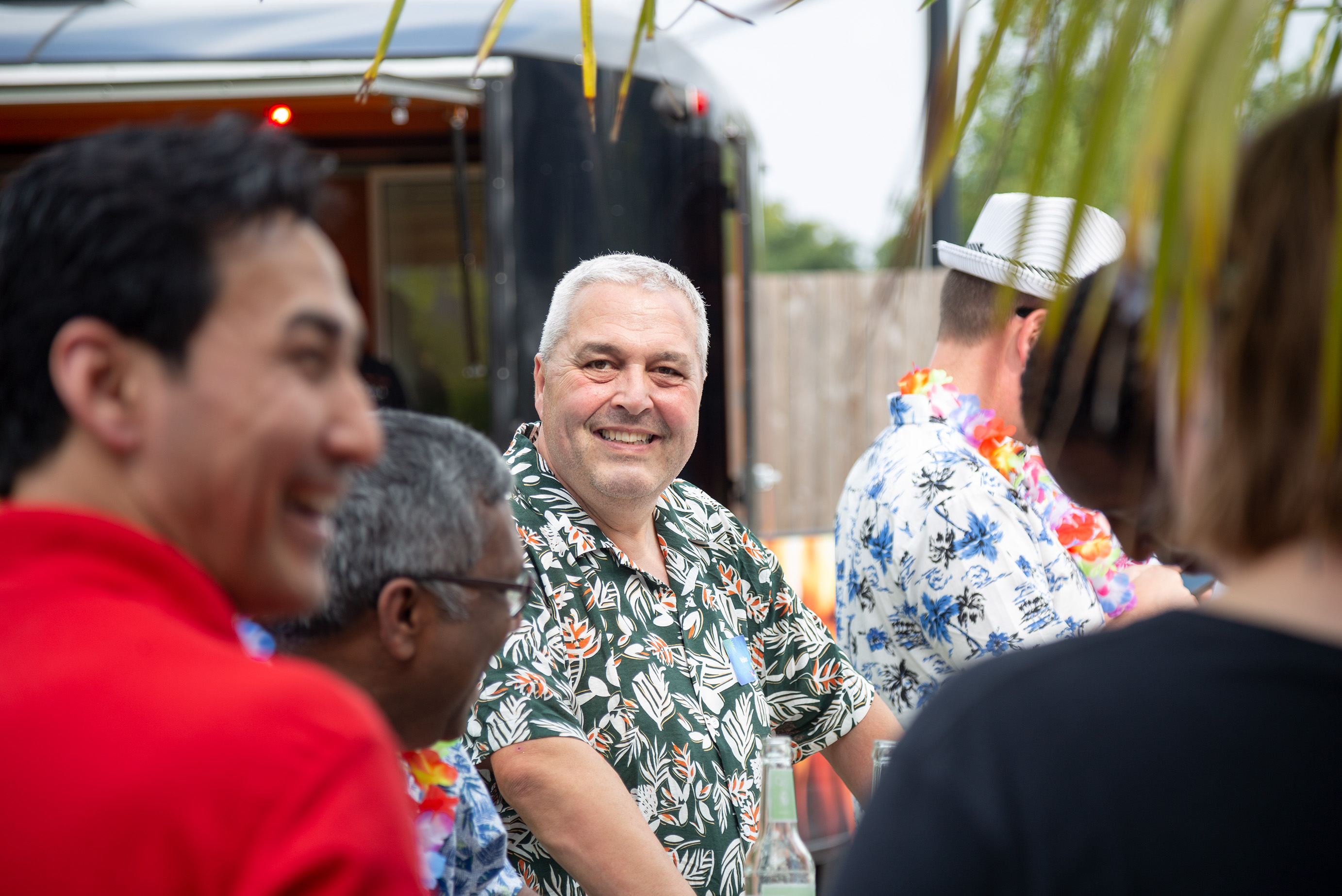 Mehrere Menschen sind von hinten zu sehen, ein Mann mit Hawaiihemd ist von vorne zu sehen und lächelt in die Kamera