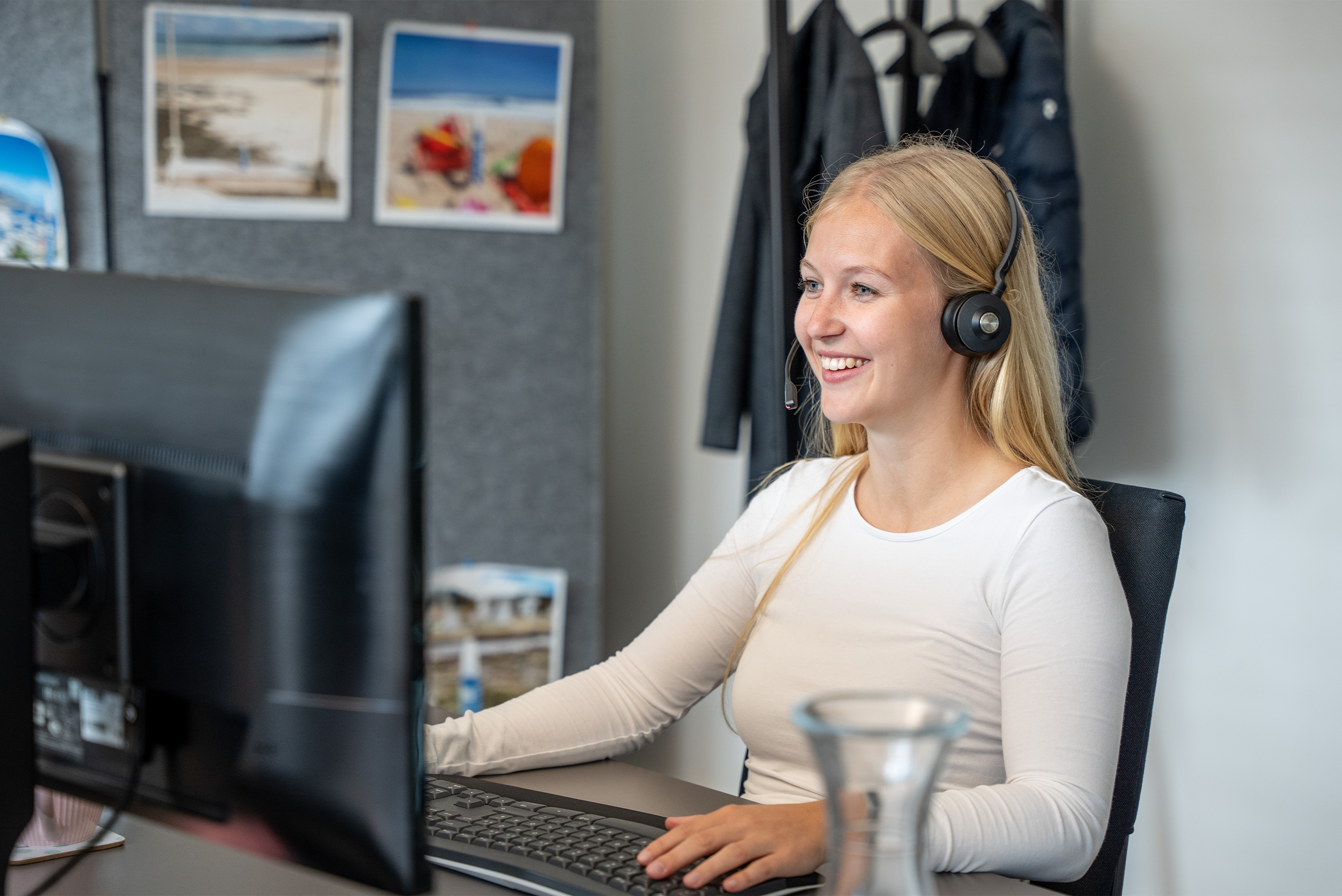 Eine junge Frau sitzt im Büro am Computer, lächelt und hat ein Headset auf